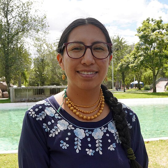 Verónica steht im Freien vor einer Wasserquelle in einer ländlichen Umgebung und blickt in die Kamera