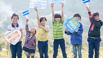 Eine Gruppe von Kindern in Vietnam hält Schilder mit Botschaften wie "I'm helping girls get equal" und "Girl power". Sie stehen auf einer Wiese und zeigen ihre Unterstützung für Geschlechtergleichstellung.