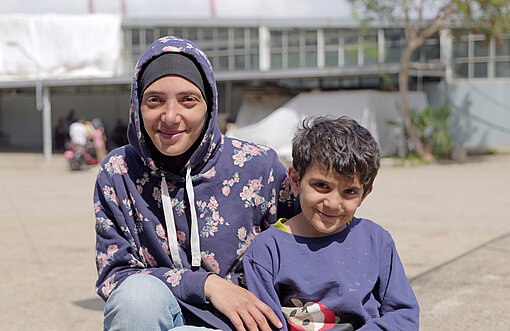 Eine Frau mit Kopftuch sitzt mit einem Kind draußen im Innenhof einer Unterkunft. Hinter ihnen ist ein helles Gebäude erkennbar.