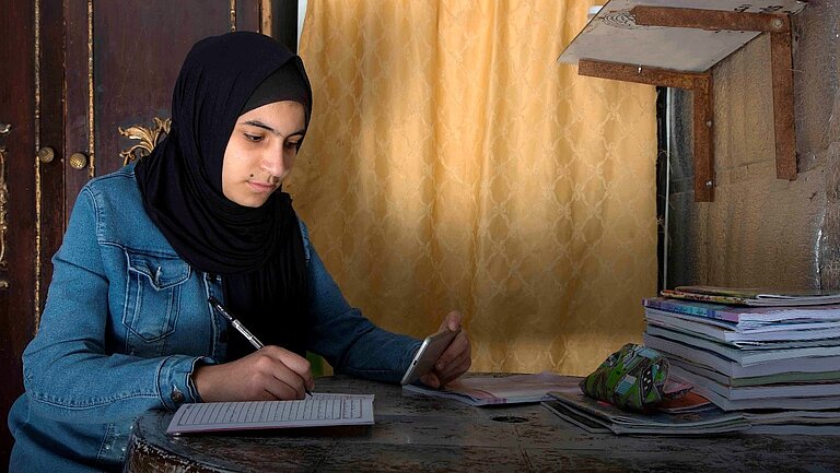 Fatima sitzt an einem Schreibtisch und schreibt auf Papier.