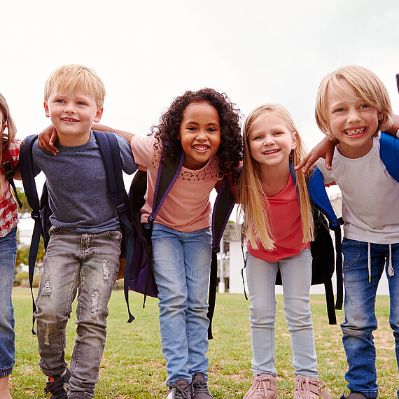 Lachende Schulkinder mit Rucksäcken beim Spielen im Freien Fröhliche Gruppe von Grundschulkindern mit Rucksäcken, Arm in Arm, im Freien