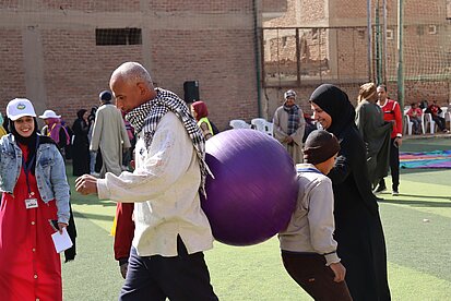 Ein Mann und ein Junge stehen mit dem Rücken zueinander. Zwischen ihren Rücken ist ein Gymnastikball eingeklemmt.
