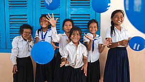 Sechs lachende Mädchen in Schuluniformen spielen mit blauen Ballons vor einem Schulgebäude mit blauen Fensterläden in Kambodscha.