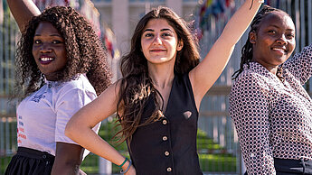 Drei selbstbewusste junge Frauen stehen mit erhobenen Armen vor dem UN-Gebäude in Genf, umgeben von zahlreichen Nationalflaggen.