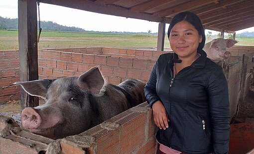 Ein Mädchen steht im Schweinestall. Sie und ihr Schwein schauen beide in die Kamera.