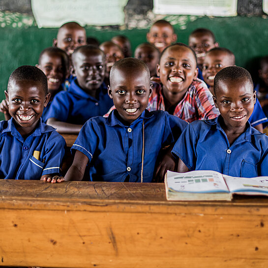 Kinder sitzen auf Schulbänken in einem Klassenzimmer und lächeln in die Kamera.