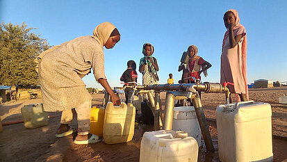 Mehrere Kinder füllen an einer niedrigen Wasserleitung Kunststoffkanister mit Wasser; die Kanister stehen auf sandigem Boden im Freien.