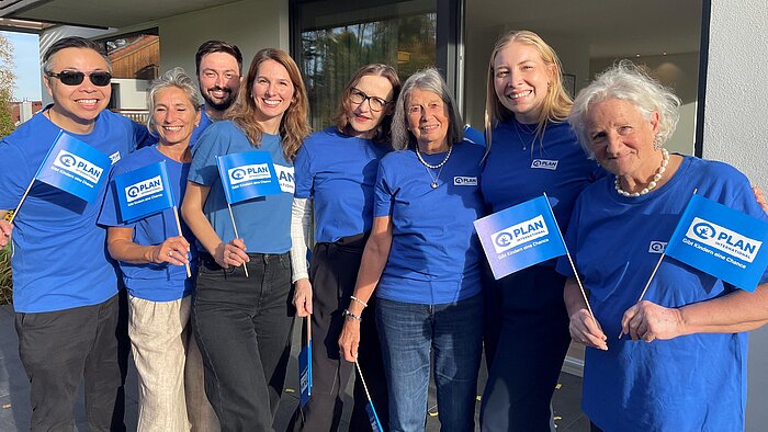 Mehrere Personen in blauen Plan-T-Shirts stehen nebeneinander und freuen sich. Sie halten blaue Fahnen mit Plan-Logo hoch.