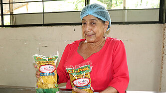 Eine Frau in einer bunten Bluse hält zwei Tüten mit Bananenchips in den Händen. Sie trägt ein Haarnetz.