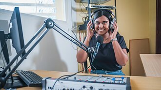 Edith sitzt an einem Radio-Mischpult, spricht in ein Mikrofon und moderiert eine Sendung für Jugendliche. Alternative Bildunterschrift: