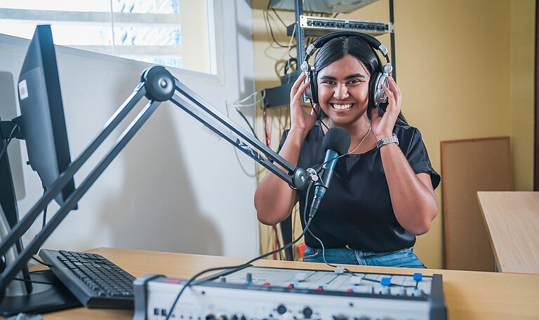 Edith sitzt an einem Radio-Mischpult, spricht in ein Mikrofon und moderiert eine Sendung für Jugendliche. Alternative Bildunterschrift: