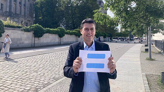 Sascha Raabe hält ein Schild hoch, auf dem das blaue Girls Get Equal-Gleichzeichen zu sehen ist.
