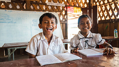 Zwei lachende Schulkinder aus Kambodscha in Schuluniform sitzen in einem Klassenraum und schreiben in ihre Hefte.
