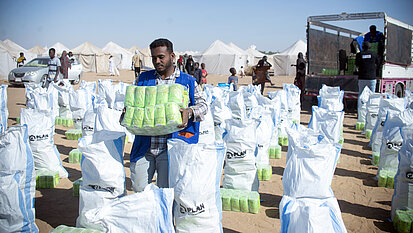 Ein Mann trägt mehrere grüne Pakete und stellt sie zwischen zahlreiche weiße Säcke; im Hintergrund stehen Zelte, ein Lastwagen und weitere Personen.