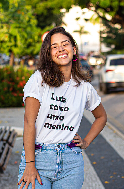 Luisa steht mit einem breiten Lächeln und einer in die Hüfte gestemmten Hand auf einem Gehweg.