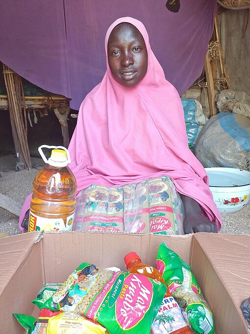 Eine afrikanische Frau in rosa Kleid sitzt vor einem Karton mit Nahrungsmitteln