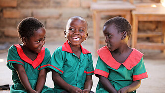 Drei lachende Kinder aus Malawi in grünen Schuluniformen mit roten Kragen sitzen nebeneinander und schauen sich fröhlich an.