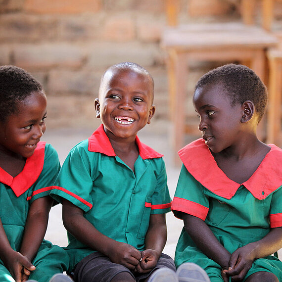 Drei lachende Kinder aus Malawi in grünen Schuluniformen mit roten Kragen sitzen nebeneinander und schauen sich fröhlich an.