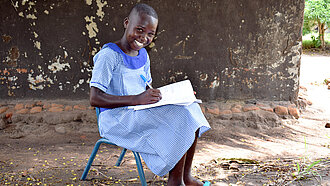 Ein Schwarzes Mädchen sitzt draußen auf einem kleinen Stuhl, schreibt etwas in ein Heft und lächelt in die Kamera
