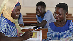 Drei ugandische Schülerinnen in Schuluniform lernen gemeinsam im Klassenraum und tauschen sich über Schulbücher aus