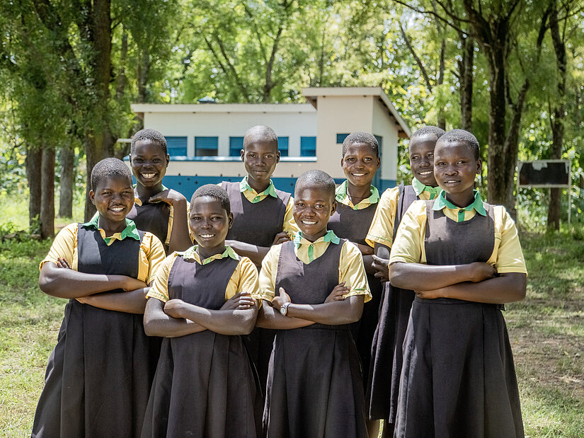 Gruppe von Schülerinnen in Schuluniformen steht gemeinsam auf dem Schulgelände vor einem Schulgebäude in Uganda