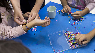 Kinder basteln bunte Armbänder aus Perlen; eine erwachsene Person hilft dabei, ein Armband am Handgelenk eines Kindes zu befestigen.