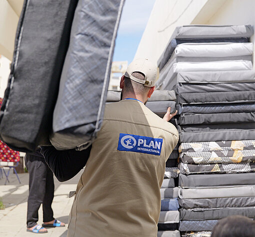 Ein Mitarbeiter von Plan International trägt Matratzen und bringt Hilfsgüter zu einer Gruppe von Menschen. Im Hintergrund ist ein Stapel Matratzen sichtbar.