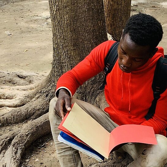 Ein Junge sitzt an einen Baum gelehnt auf dem Boden und liest in einem Schulheft