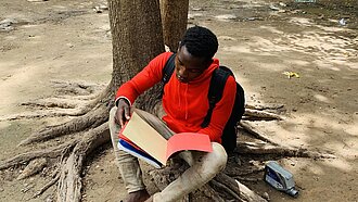 Ein Junge sitzt an einen Baum gelehnt auf dem Boden und liest in einem Schulheft