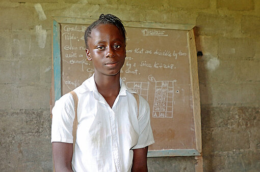 Ein Mädchen in weißer Bluse steht vor einer Schultafel