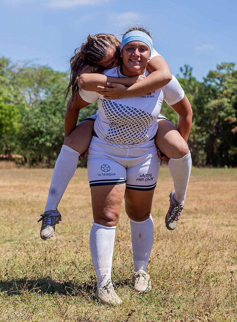 Eine junge Frau trägt eine andere junge Frau Huckepack. Beide lachen und tragen Fußball-Trikots.