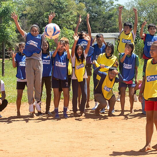 Ein Mädchen steht auf einem Sandplatz und schießt einen Ball, am Spielfeldrand stehen Jungen und Mädchen und feuern die Spielerin an