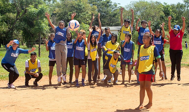Ein Mädchen steht auf einem Sandplatz und schießt einen Ball, am Spielfeldrand stehen Jungen und Mädchen und feuern die Spielerin an