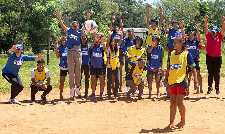 Ein Mädchen steht auf einem Sandplatz und schießt einen Ball, am Spielfeldrand stehen Jungen und Mädchen und feuern die Spielerin an