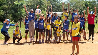 Ein Mädchen steht auf einem Sandplatz und schießt einen Ball, am Spielfeldrand stehen Jungen und Mädchen und feuern die Spielerin an