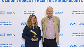 Eine Frau und ein älterer Mann stehen vor einer Pressewand. Die Frau hält stolz einen Preis in den Händen