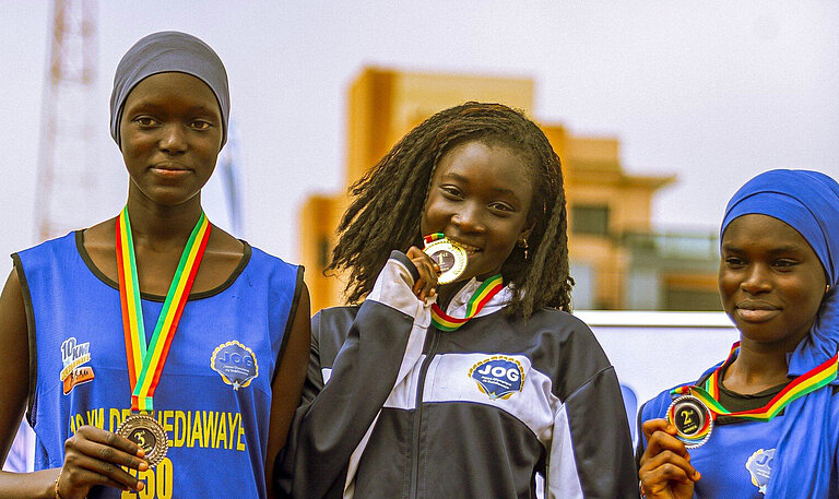 Drei junge Senegalesinnen stehen auf einem Podest, jede hat eine Medaille umhängen. Das Mädchen in der Mitte beißt auf ihre Medaille.