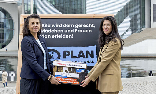 Zwei Frauen in schicker Kleidung stehen in Berlin vor dem Bundestag, im Hintergrund ist der Fluss Spree und das Plan-Logo zu sehen
