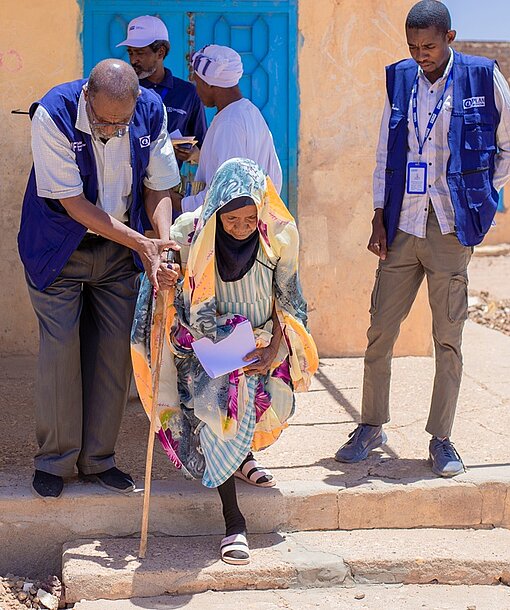 Ein Mitarbeiter von Plan International hilft einer älteren Frau eine Treppe herunter