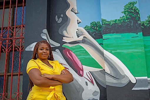 Eine dunkelhäutige Frau in leuchtend gelbem Kostüm steht vor einer Wand mit einem großen Graffitibild.