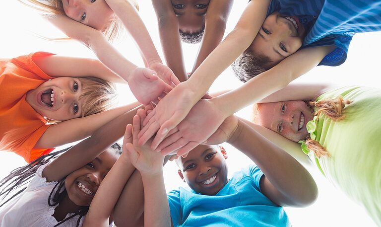  Mehrere Kinder bilden einen Kreis und legen lächelnd ihre Hände zur Mitte zusammen