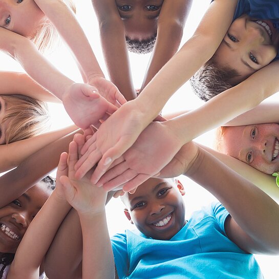  Mehrere Kinder bilden einen Kreis und legen lächelnd ihre Hände zur Mitte zusammen