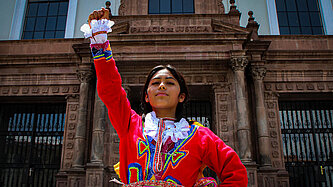 Ein Mädchen in traditioneller Kleidung steht vor einem Gebäude und reckt eine Raust in die Luft