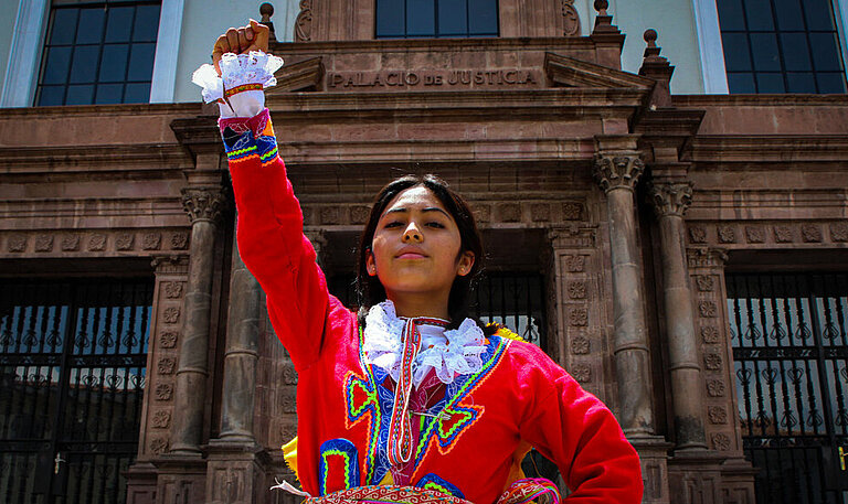 Ein Mädchen in traditioneller Kleidung steht vor einem Gebäude und reckt eine Raust in die Luft