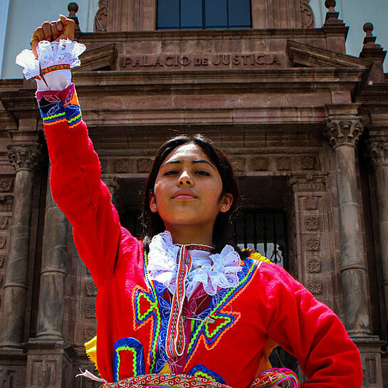 Ein Mädchen in traditioneller Kleidung steht vor einem Gebäude und reckt eine Raust in die Luft