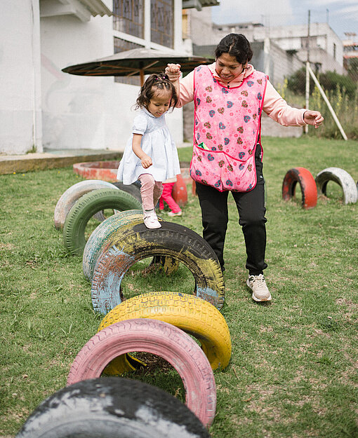 Eine Frau unterstützt ein Kind beim Ausbalancieren auf alten Reifen in einem Außenbereich. Beide lächeln, während sie gemeinsam spielen.