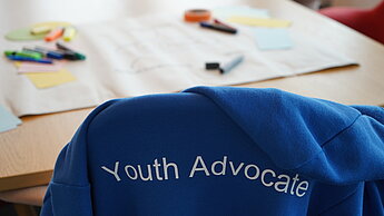 Im Vordergrund hängt über einem Stuhl ein blauer Kapuzenpullover mit der Aufschrift "Youth Advocate". Der Stuhl steht an einem Tisch auf dem Papier und verschiedene Stifte liegen.