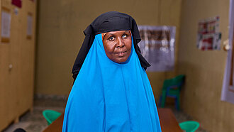Eine Frau in leuchtend blauem Gewand und schwarzem Kopftuch schaut in die Kamera. Sie steht vor einem Konferenztisch.