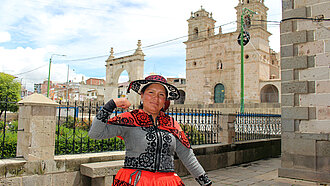 Jessica aus Chumbivilcas, Peru, steht vor einer historischen Kirche und zeigt stolz ihre Stärke, während sie traditionelle Kleidung trägt.