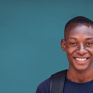 Ein junger Mann steht vor einem blau-grünen Hintergrund und lächelt in die Kamera.
